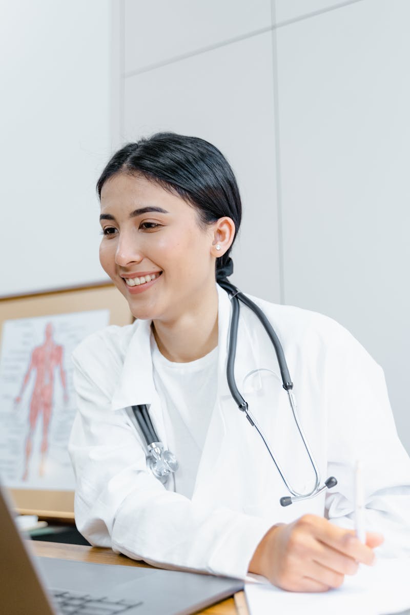Confident female doctor in a lab coat smiling during an online consultation using a laptop.